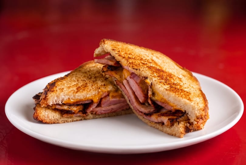 Grilled ham and cheese sandwich, cut in half, on a white plate against a red background.