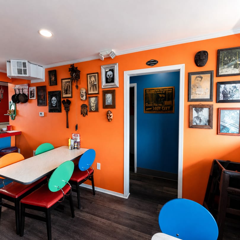 A vibrant orange restaurant interior with eclectic wall decor, colorful chairs, and a doorway to restrooms.