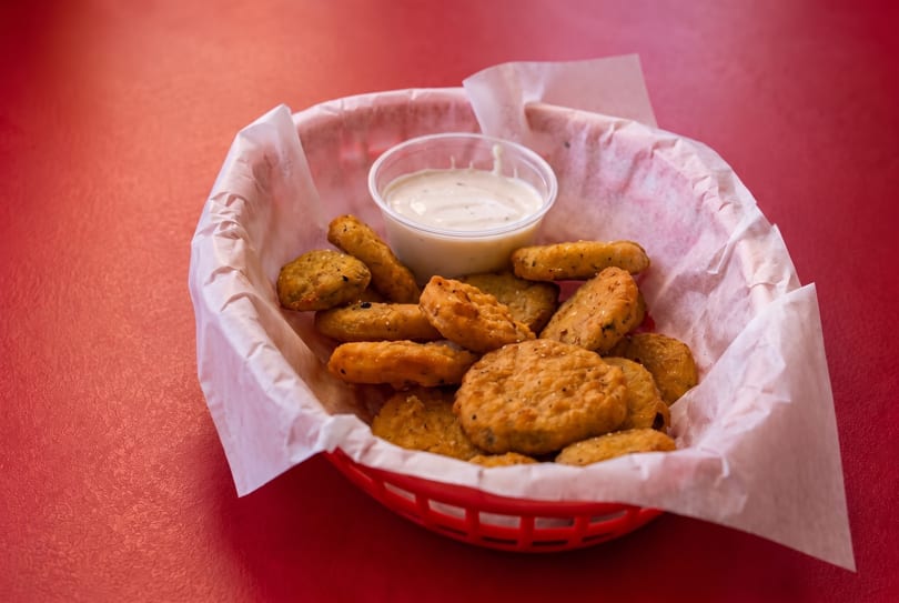 A red plastic basket filled with crispy fried pickles and a side of ranch dressing on a red table.