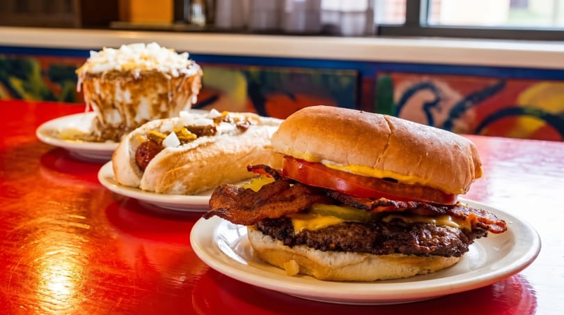 A bacon cheeseburger, hot dog, and chili cheese fries on a red table.