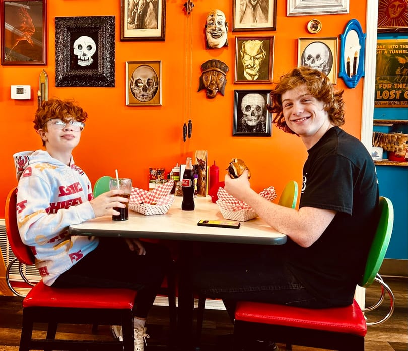 Two boys sit at a diner table with burgers and drinks, against an orange wall with skull art.